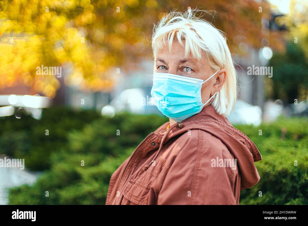 Kaukasische Erwachsene blonde Frau 50 Jahre alt in heller Robe und schützende medizinische Atemschutzmaske Blick auf den Park Hintergrund Stockfoto