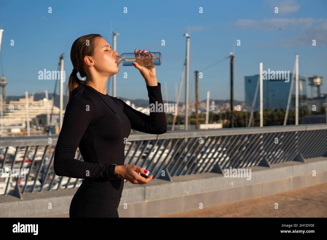 Weibliche Athletin mit Pferdeschwanz schlürft während des Fitnesstrainings am Ufer in der Nähe des Hafens in der Stadt frisches Wasser aus der Flasche. Stockfoto