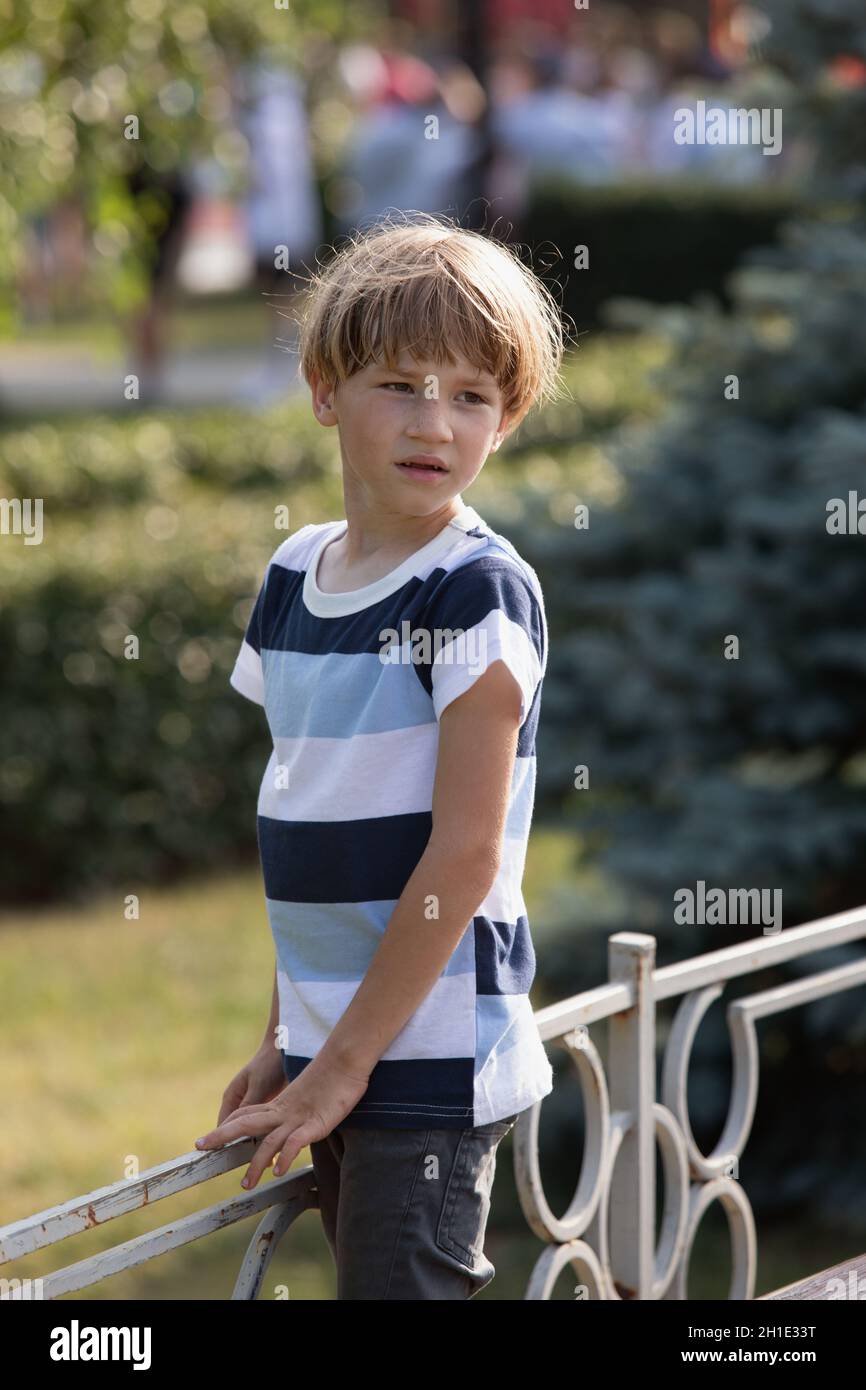 Kleiner, blonder Junge in gestreiftem Hemd und Jeans am sonnigen Sommermorgen. Grüne Bäume im Hintergrund. Konzentration in jungen Augen. Stockfoto
