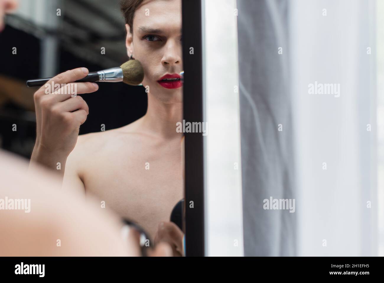 Junger Transgender-Mann, der Gesichtspuder mit einem Kosmetikpinsel in der Nähe des Spiegels anwendet Stockfoto