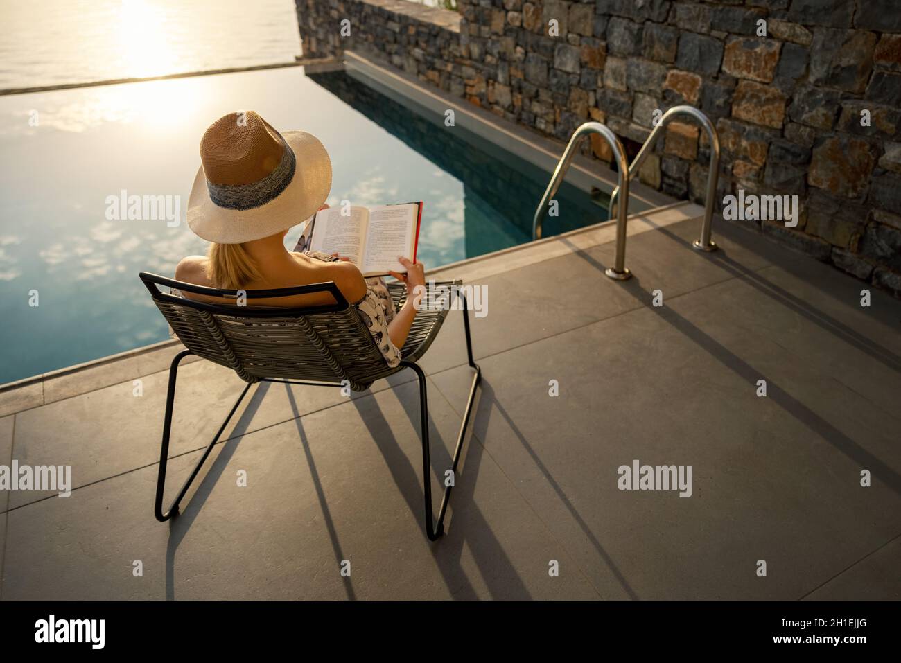 Erholsamer Urlaub - Frau sitzt im Stuhl und liest ein Buch auf der Terrasse in der Nähe des Swimmingpools in der Luxusvilla Stockfoto