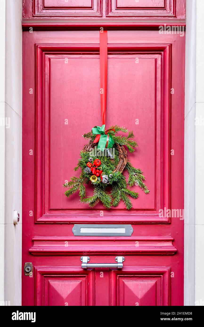 Vorderansicht einer Weihnachtskrone aus Tannenzweigen, Tannenzapfen und Bändern, die an der roten Haustür mit Zierleisten eines Stadthauses hängt. Stockfoto