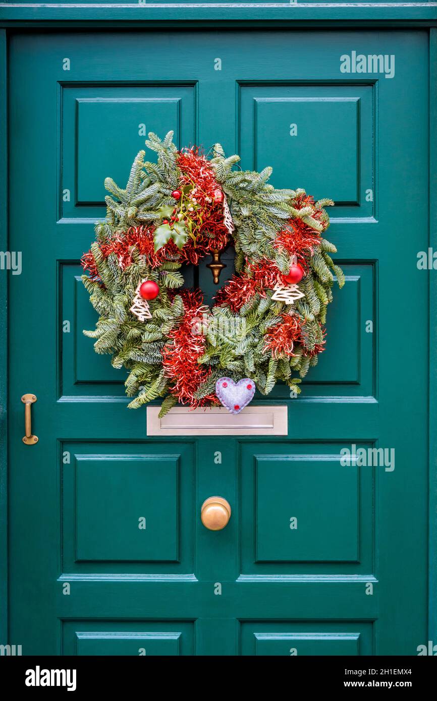 Vorderansicht einer Weihnachtskrone aus Tannenzweigen, Lametta und Kugeln, die an einer grünen Haustür mit Zierleisten hängt. Stockfoto