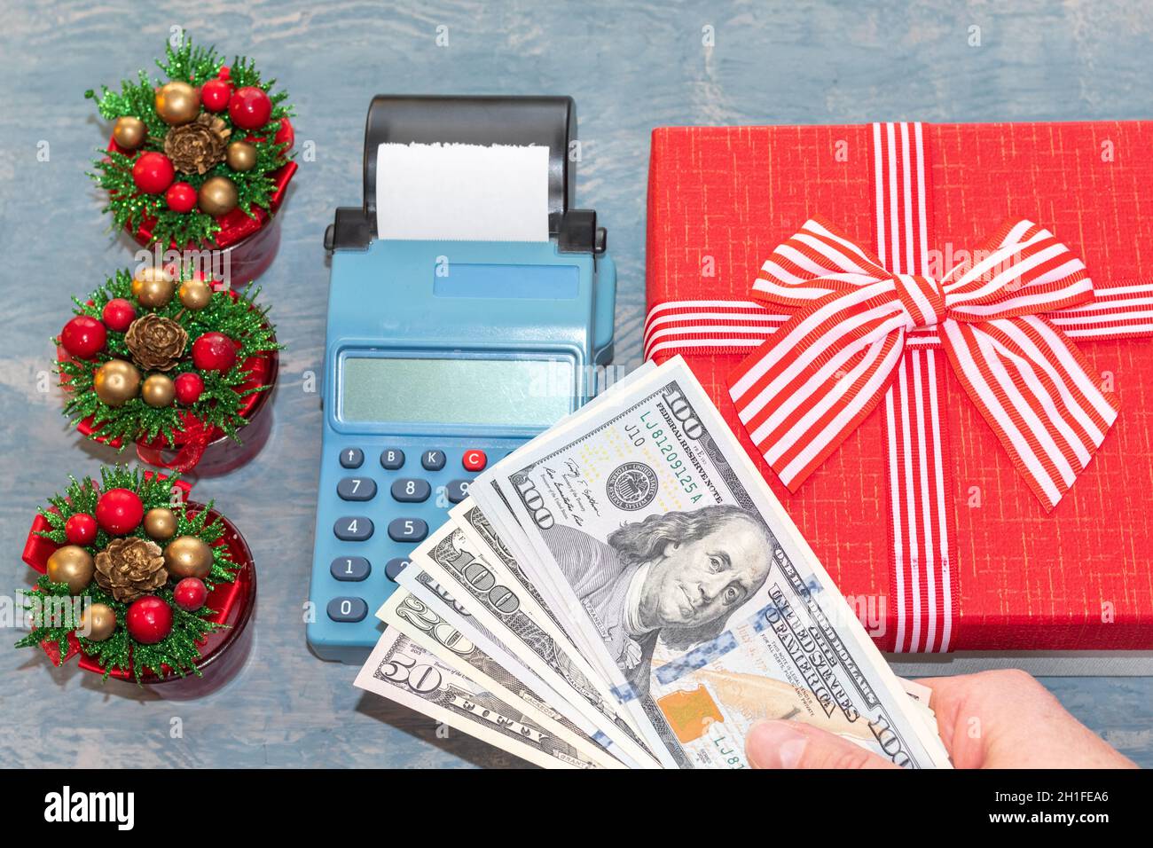 Weihnachtsangebot. Eine Hand mit Dollars über einer Kasse in der Nähe einer roten Geschenkbox mit einem Band und kleinen Weihnachtsbäumen mit Zapfen. Online-Shopping. B Stockfoto