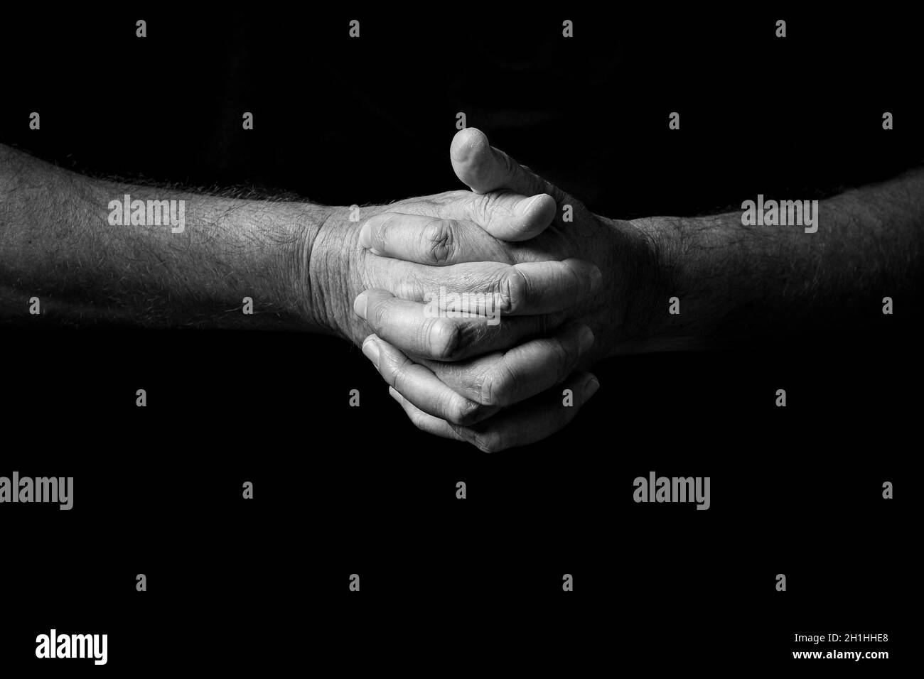 Schwarz-weißes Bild von zerknitterten männlichen Händen, die in Gedanken oder im Gebet zusammengehalten und vor einem schwarzen Hintergrund mit dramatischem Licht isoliert wurden Stockfoto