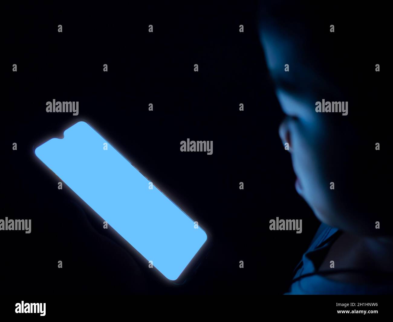 Ein Junge benutzt ein Telefon mit einem leeren blauen Bildschirm auf einem dunkelschwarzen Hintergrund. Stockfoto