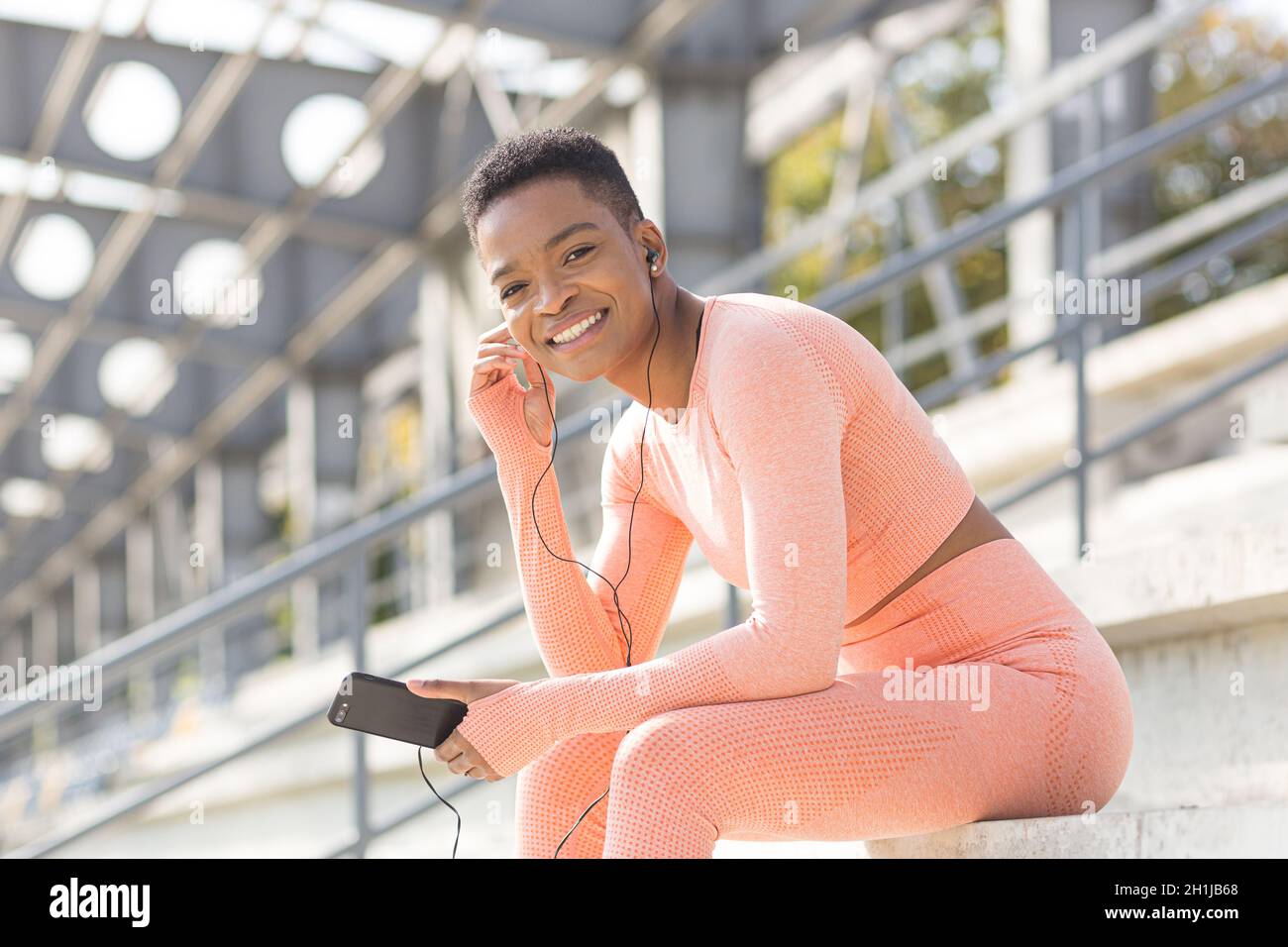 Junge schöne Frau in einem rosa Sportanzug lächelt und schaut auf die Kamera, nach dem Training hört Musik mit Kopfhörern und einer Anwendung auf einem m Stockfoto