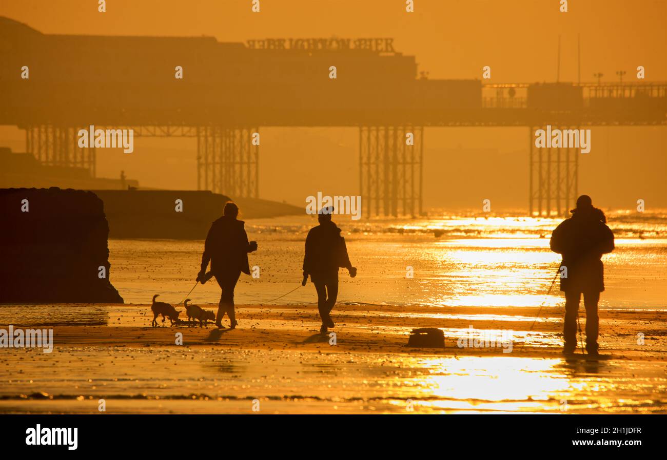 Brighton Beach bei Ebbe, Sonnenaufgang. Der Palace Pier im Hintergrund. Frauen gehen mit Hunden. Brighton & Hove, Sussex, England, Großbritannien Stockfoto