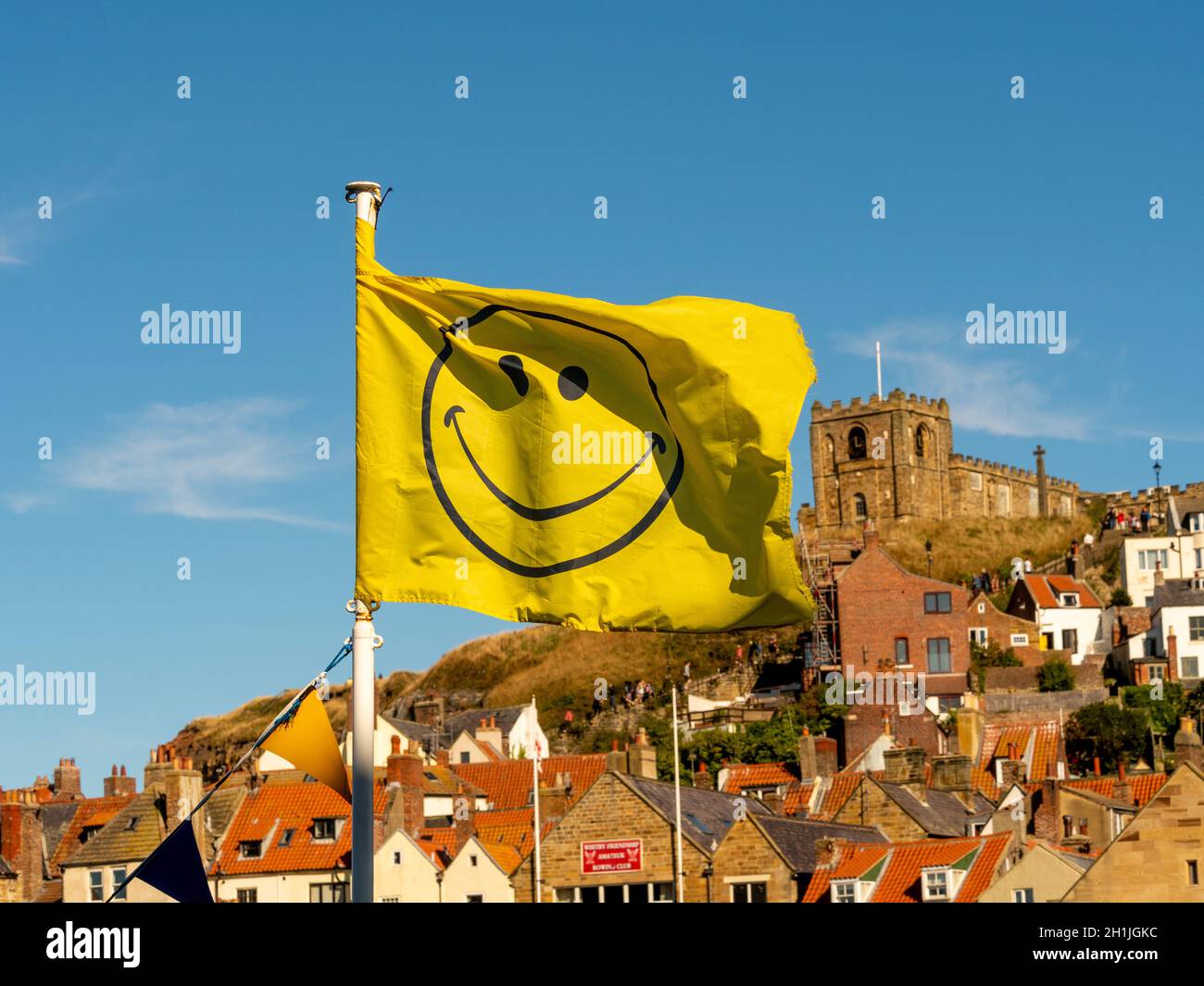 Gelbe Smiley-Flagge flattert im Wind an einem sonnigen Tag mit der St. Mary's Church, Whitby in der Ferne. North Yorkshire, Großbritannien Stockfoto