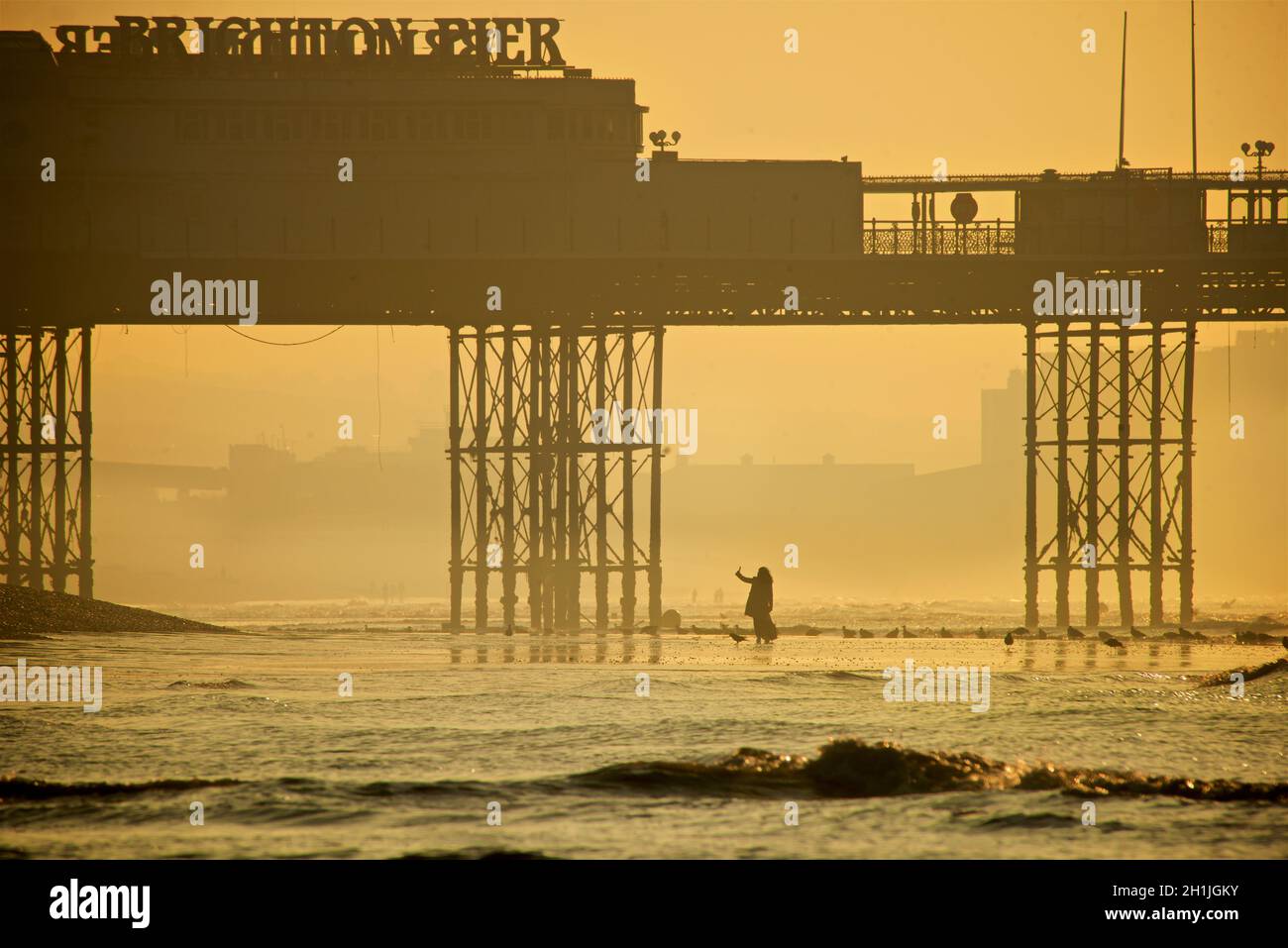 Brighton Beach bei Ebbe, Sonnenaufgang. Der Palace Pier im Hintergrund. Silhouetten von Frauen im langen Mantel, die Selfies machen Brighton & Hove, Sussex, England, Großbritannien Stockfoto