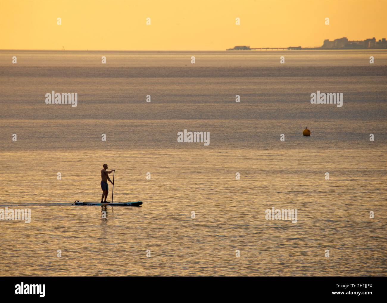 Ein silhouettierter Paddlebarder, der bei Sonnenuntergang an einem Hove Strand paddeln kann. Brighton & Hove, Engand, Großbritannien. Worthing Pier, West Sussex am Horizont. Stockfoto