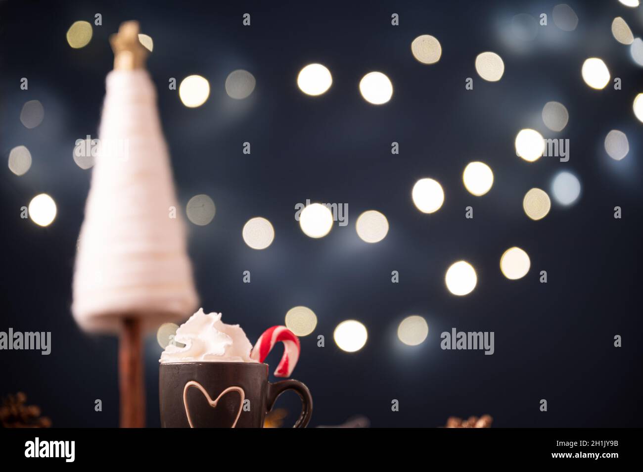 Kleine Tasse heiße Schokolade mit Schlagsahne verziert mit Zuckerrohr neben einer weihnachtsdekoration und verschwommene Weihnachtslichter im Hintergrund. Stockfoto