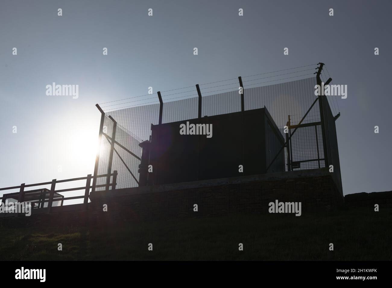 Hohe Stacheldraht Anti klettern Zaun umhüllt große quadratische Metall industriellen Wassermanagement Ausrüstung. Blick nach oben, blauer Himmel und helle Sonne dahinter Stockfoto