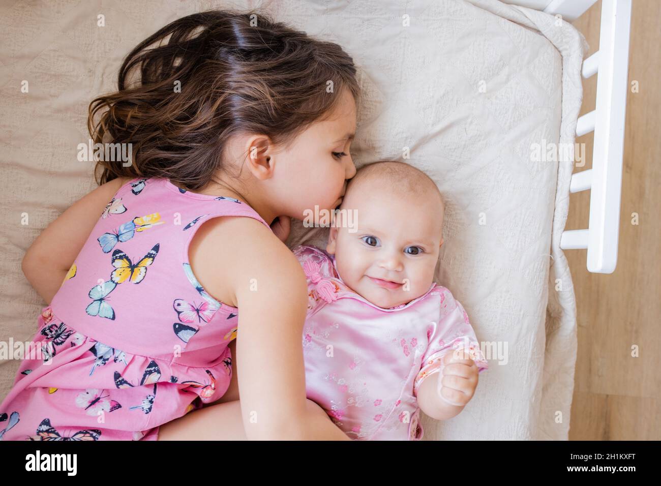 Das kleine Mädchen liegt liebevoll und friedlich auf dem Bett und umarmt und küsst ihre glückliche kleine Schwester. Niedliche Schwestern in rosa Mäntel, die auf einem Bett ruhen. Junge Sibli Stockfoto