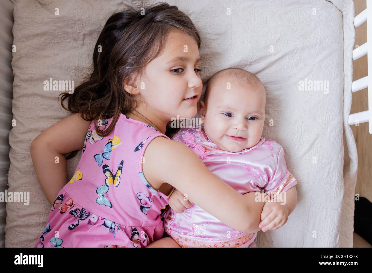 Glückliches kleines Mädchen liebevoll umarmt Baby Schwester in rosa  asiatische Kleidung auf einem Bett. Zwei liebliche Geschwister in rosa  Mäntel liegen auf dem Bett. Junge Geschwister ha Stockfotografie - Alamy