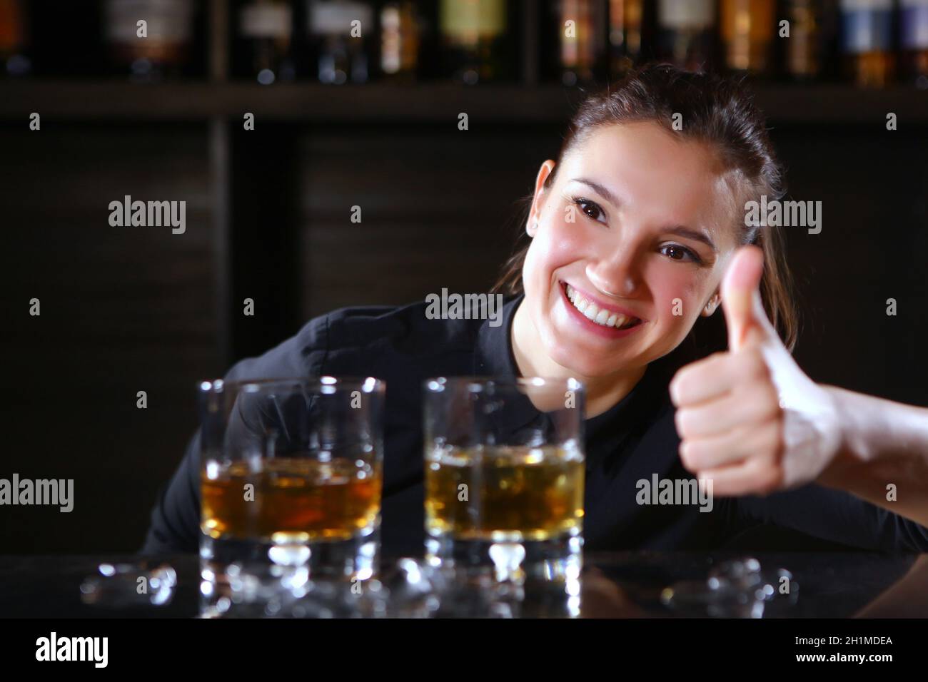 Das Barkeeper-Mädchen lächelt und hält ihren Daumen hoch. Foto im Inneren einer Bar in einem Restaurant. Foto auf schwarzem Hintergrund. Speicherplatz kopieren. Stockfoto