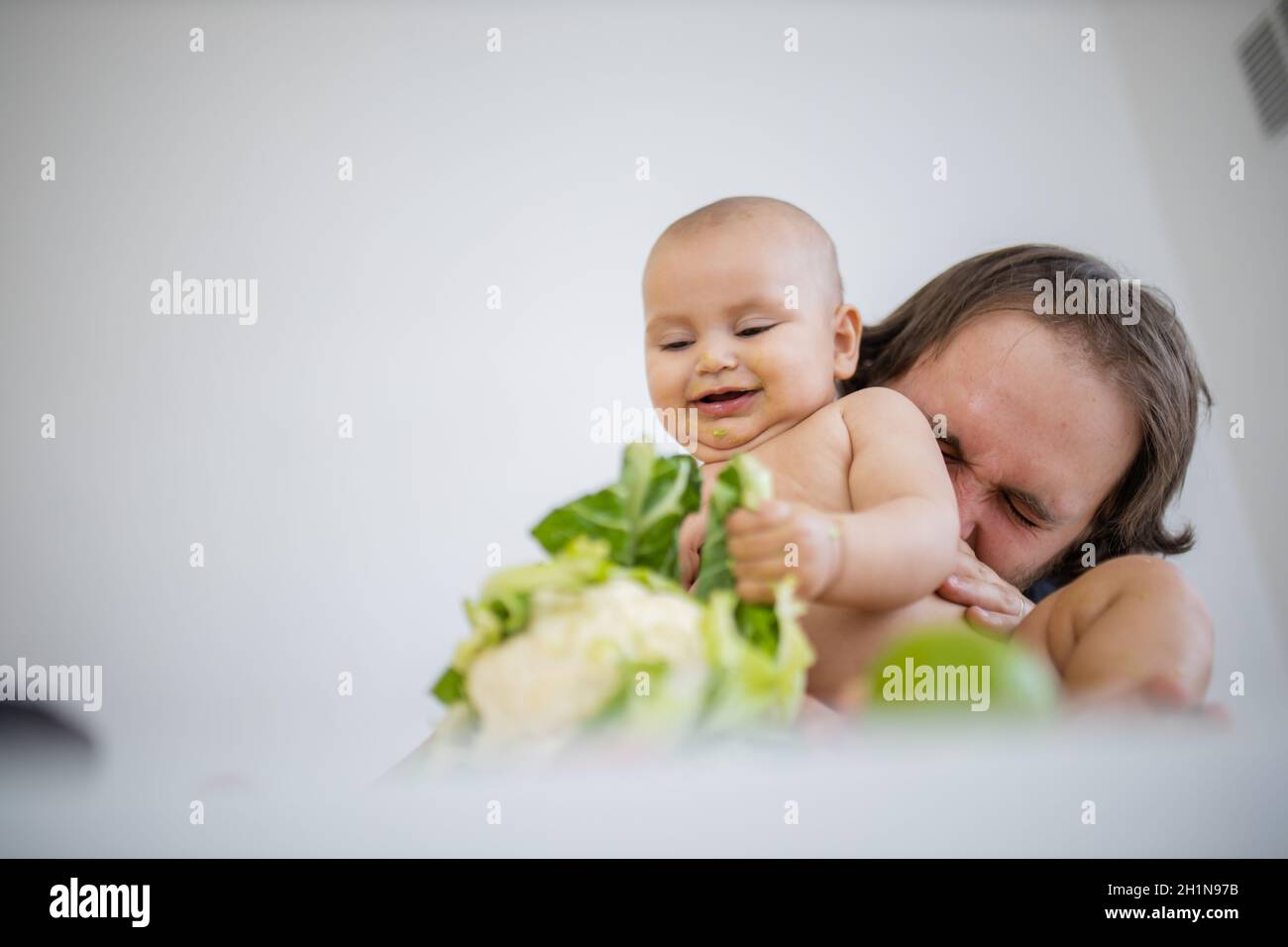 Vater hält und küsst seine glückliche Tochter liebevoll über dem Tisch. Entzückendes Baby lächelt und spielt mit Blumenkohl. Babys interagieren mit f Stockfoto