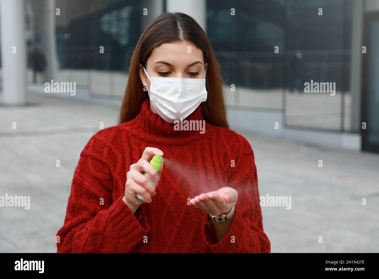 Portrait der jungen Frau trägt schützende medizinische Maske Sprühen Alkohol Desinfektionsmittel an den Händen in der modernen Stadtstraße Stockfoto