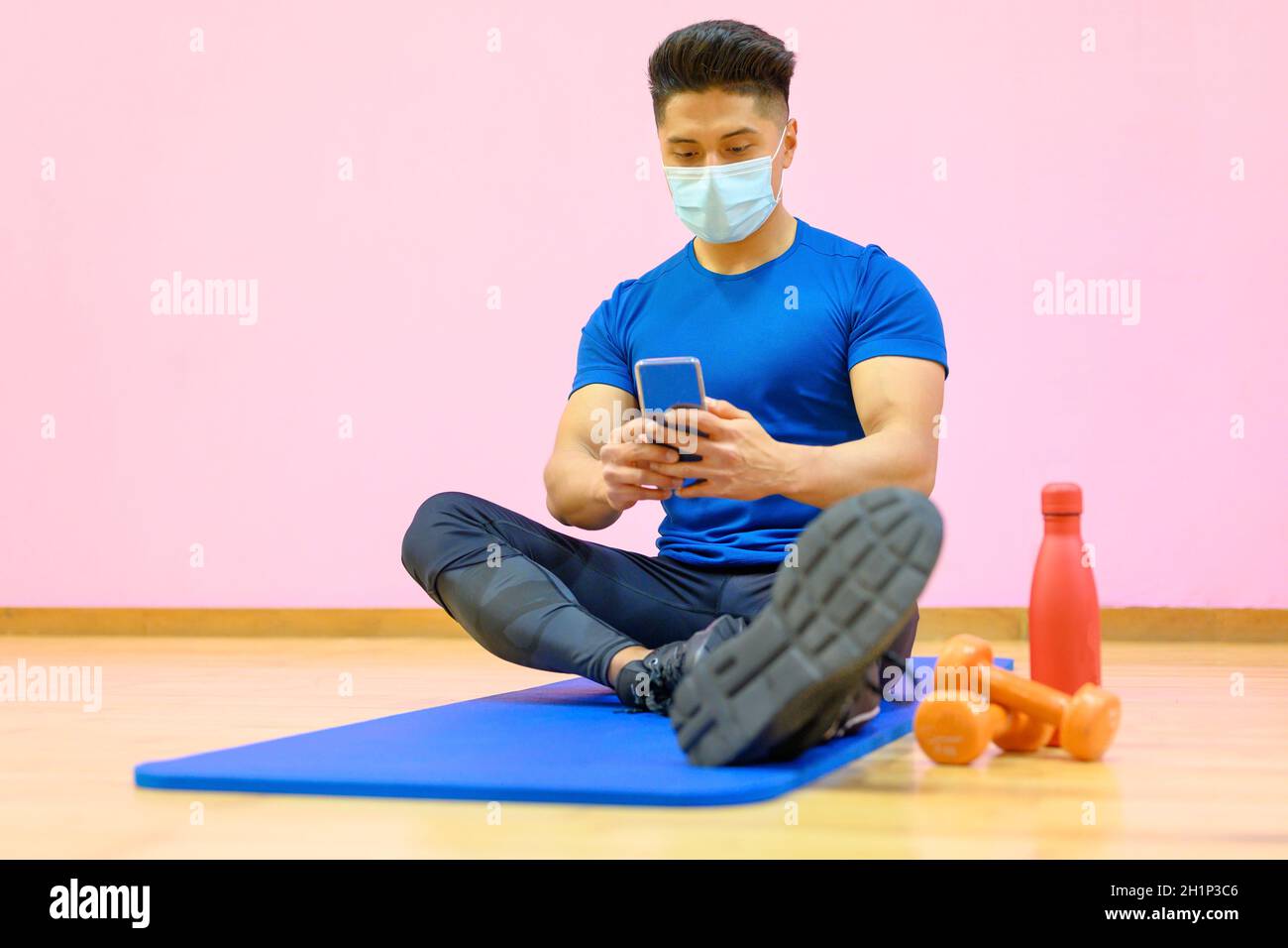 Junger Latein-Mann mit Schutzmaske, entspannt, mit Handy nach dem Training im Fitnessstudio während Pandemie neue Normalität. Hochwertige Fotos Stockfoto