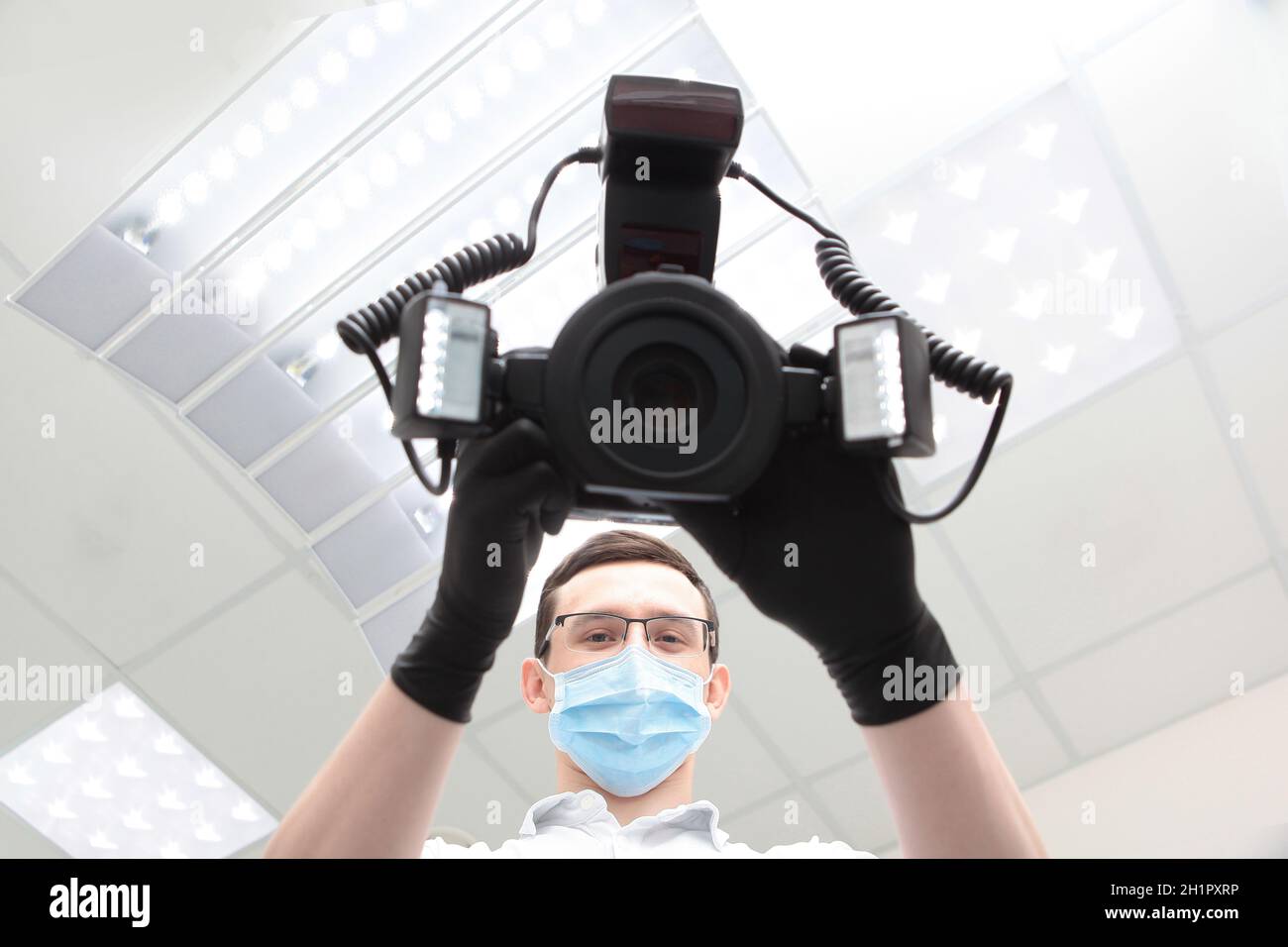 Eine Kamera mit einem Ringblitz in den Händen eines Arztes. Arzt in einer medizinischen Maske. Hände in schwarzen Schutzhandschuhen. Moderne Zahnarztpraxis. Ansicht von unten. TC Stockfoto