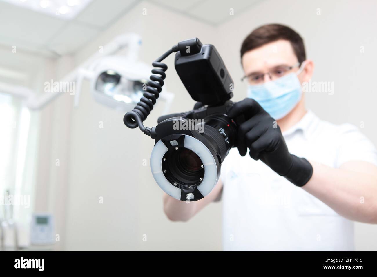 Der Zahnarzt macht während des Termins ein Foto auf der Kamera. Arzt in Schutzmaske außer Fokus. Eine Kamera mit einem Ringblitz in den Händen eines Stockfoto
