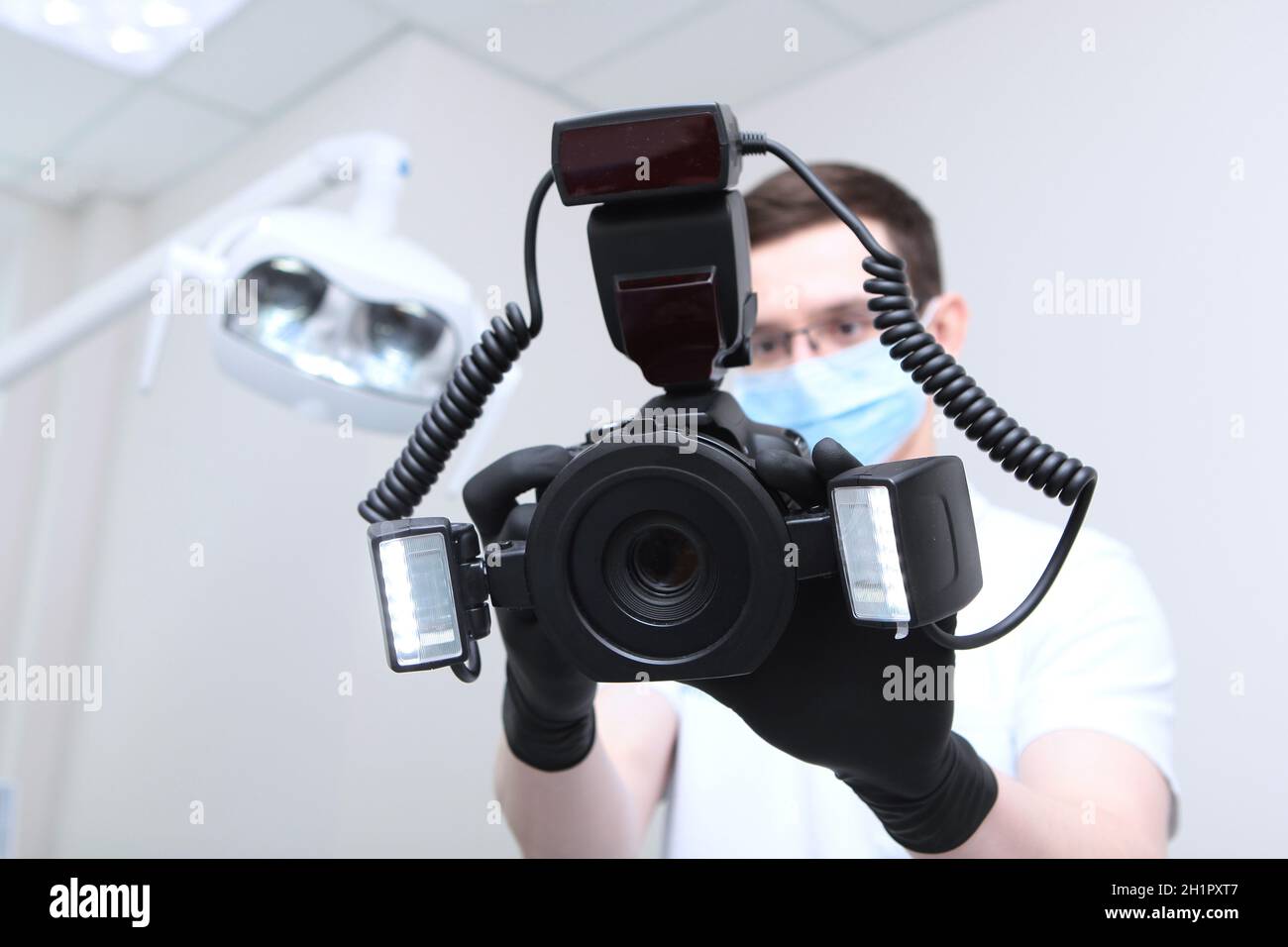 Der Zahnarztarzt macht ein Foto mit einer Kamera. Eine Kamera mit einem Ringblitz in den Händen eines Arztes. Arzt in einer medizinischen Maske. Hände in schützendem Schwarz Stockfoto