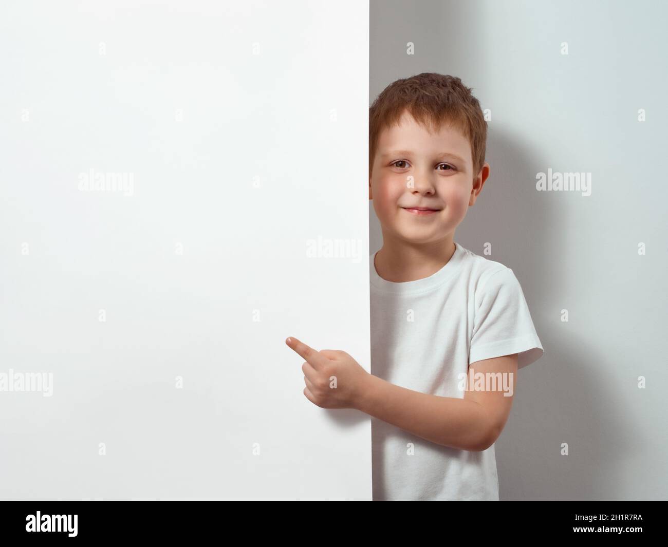 Der vierjährige Junge zeigt Finger auf einem weißen Blatt. Happy Child in weißem T-Shirt auf weißem Hintergrund mit Kopieraum für Nachricht, Modell Stockfoto