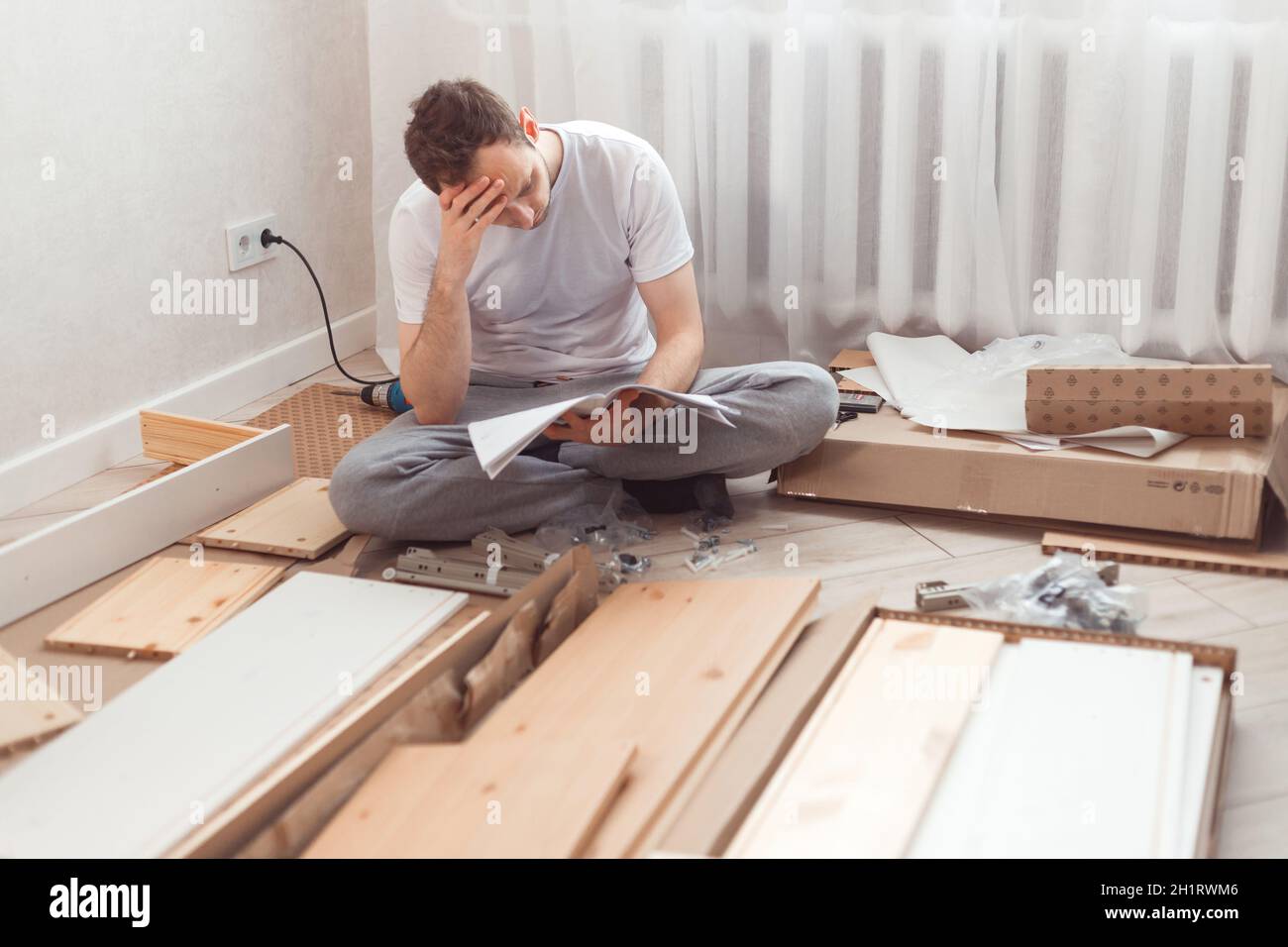 Verwirrter Mann, der Holzmöbel zusammengebaut hat. Schwierigkeiten und Nachteile der Selbstmontage der Möbel zu Hause ohne Meister. Mann beim Lesen von Anweisungen Stockfoto
