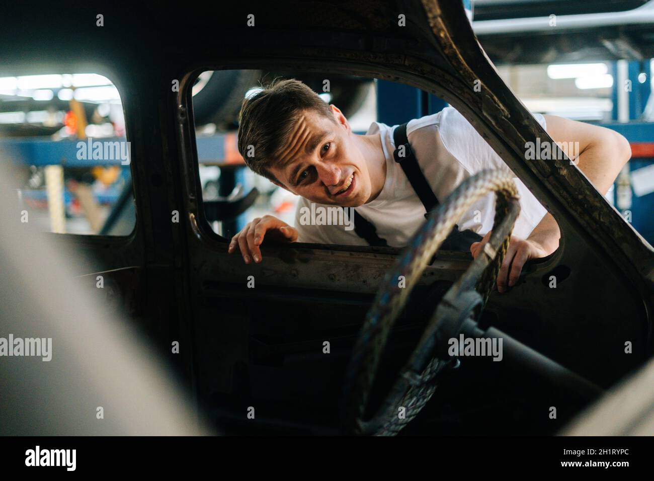 Vorderansicht des frustrierten jungen Service-Mannes in Uniform, der das Innere des alten Autos in der Autowerkstatt mit Fahrzeughintergrund inspiziert. Stockfoto