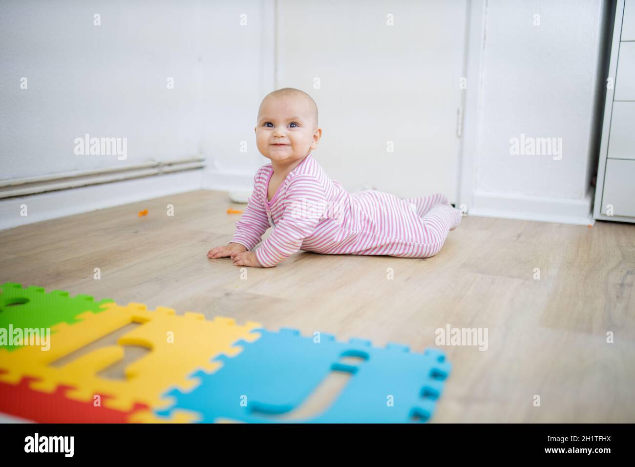 Entzückendes Baby lächelt und liegt mit dem Gesicht nach unten auf dem Holzboden neben der farbenfrohen Kindermatte. Porträt von niedlichen Baby spielen auf dem Boden. Glückliche Babys havi Stockfoto