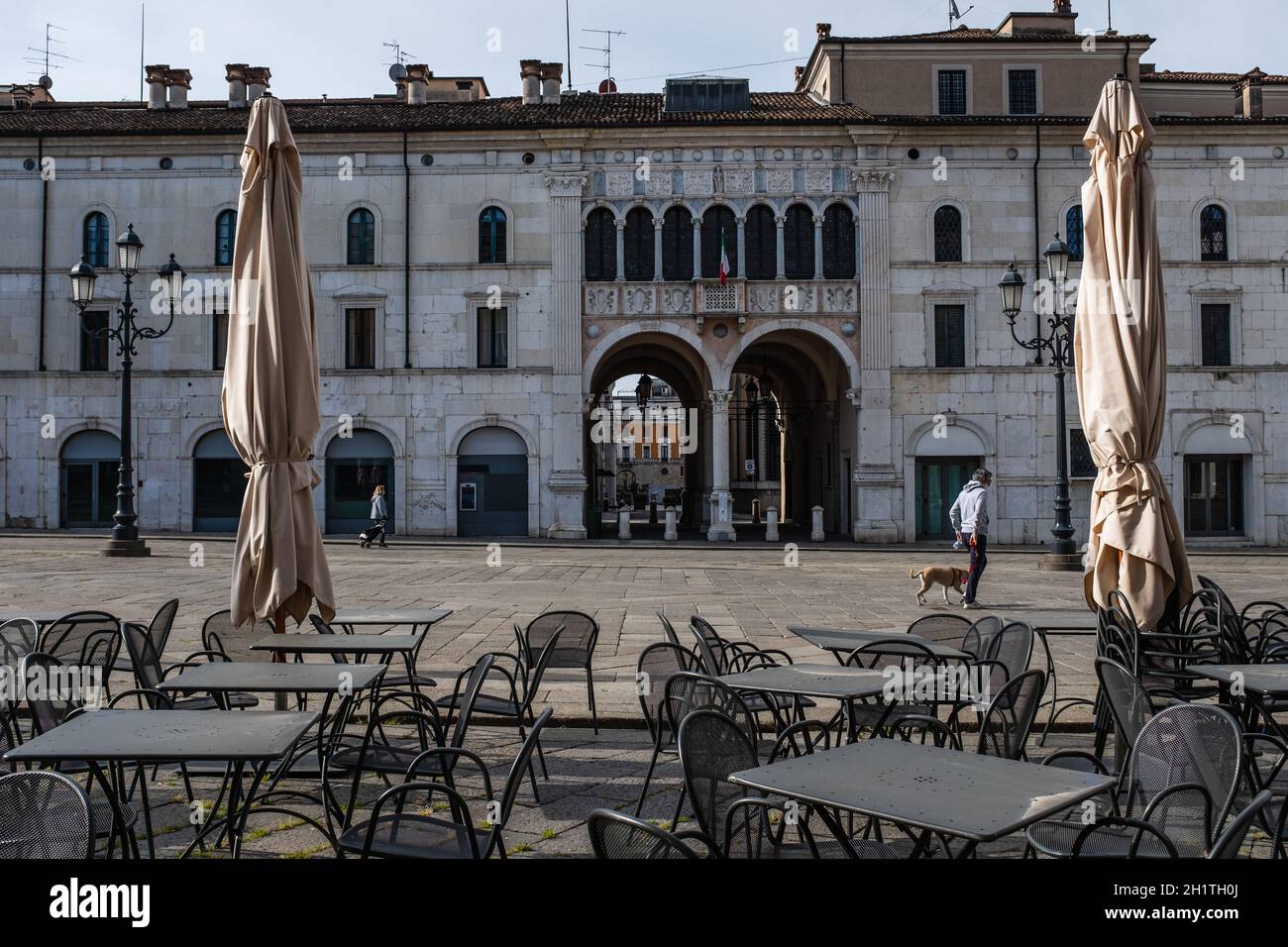 Menschenleere Tische und Stühle vor dem Restaurant. Nur zwei Personen, die während der Coronavirus-Notverriegelung über den Hauptplatz von Brescia laufen Stockfoto