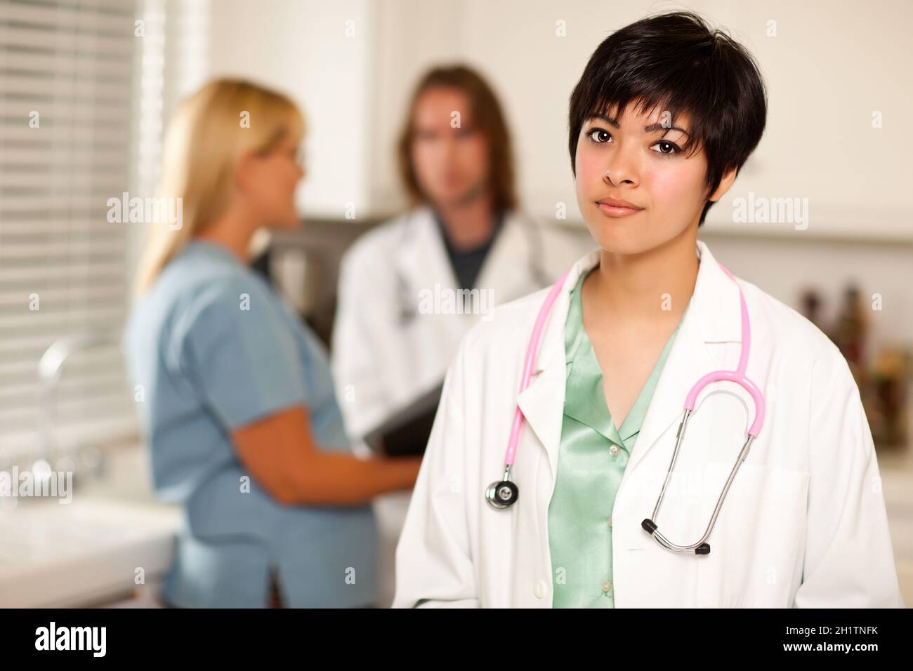 Der hübsche Latino-Arzt schlint an der Kamera, während Kollegen hinter ihr reden. Stockfoto