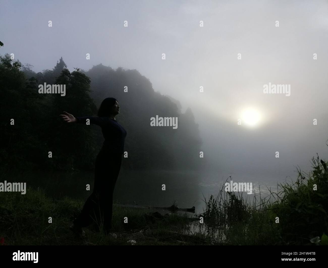 Junge asiatische Frau, die allein auf dem See steht und einen friedlichen Moment des Sonnenaufgangs in grau-bewölktem Himmel am frühen, kühlen Morgen genießt. Konzepte des Gewinners, Stockfoto