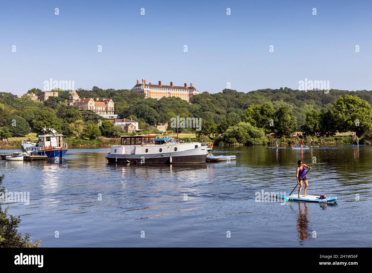 Ein Paddelboarder fährt auf der Themse in Meadowside, East Twickenham, mit dem Petersham Hotel und den Royal Star & Garter Home Buildings dahinter. Stockfoto