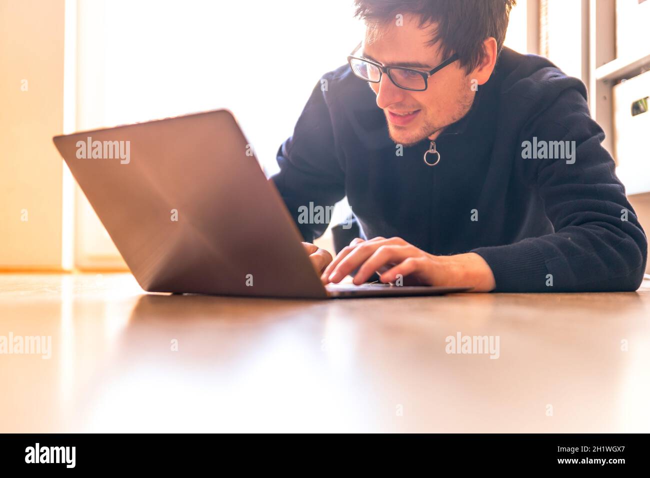 Glückliche junge Mann mit einem Laptop arbeitet auf dem Holzboden, Licht aus dem Fenster kommt Stockfoto