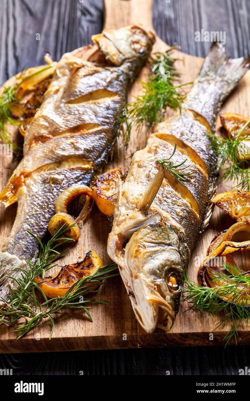 Gebratener Seebarsch-Fisch auf einem Holzbrett mit Meersalz, Dill, Zitrone auf einem schwarzen Holztisch, Draufsicht, Nahaufnahme Stockfoto