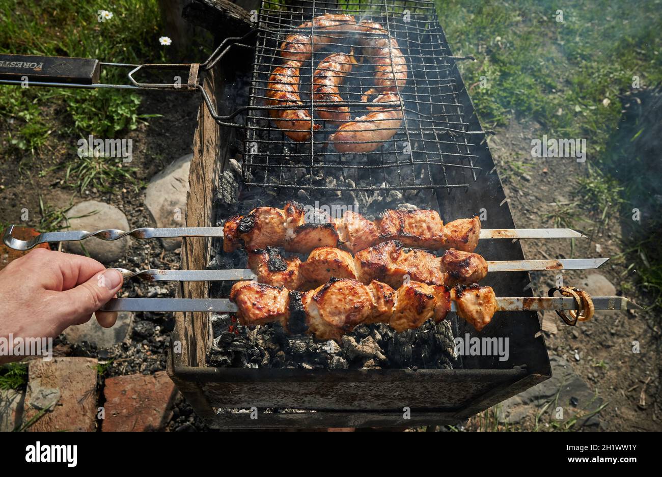 Mann, der Schaschlik oder Schischkebab über Holzkohle vorbereitet. Grillen Sie Fleisch und Würstchen oder Bratwurst auf einem Grillrost im Hinterhof. Gegrilltes Fleisch auf Metallspieß Stockfoto