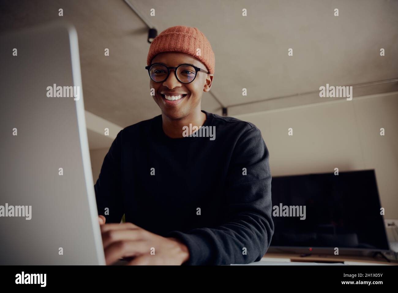 Schwarzer Mann lächelt auf die Kamera, während er auf dem Laptop an der Küchentheke sitzt. Junge Erwachsene arbeiten von zu Hause aus Stockfoto