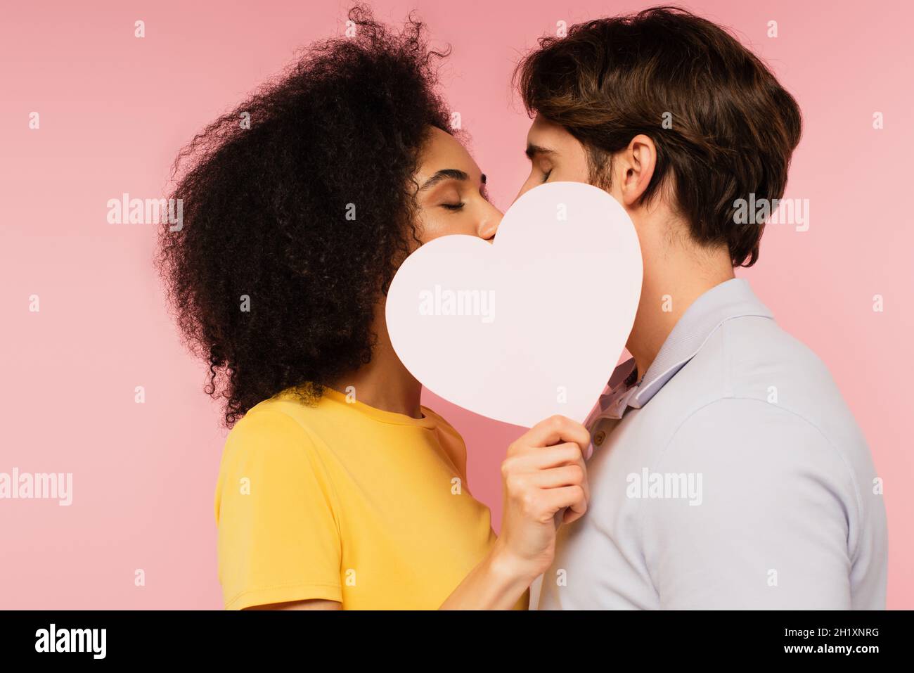 Interracial paar mit geschlossenen Augen küssen hinter leerem Papier Herz isoliert auf rosa Stockfoto