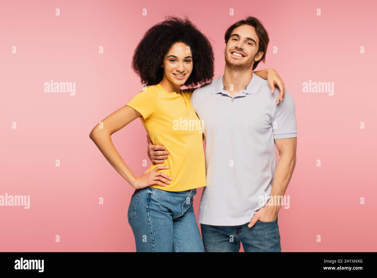 Junges und fröhliches interrassisches Paar, das die Kamera anschaut, während es isoliert auf Pink umarmt Stockfoto