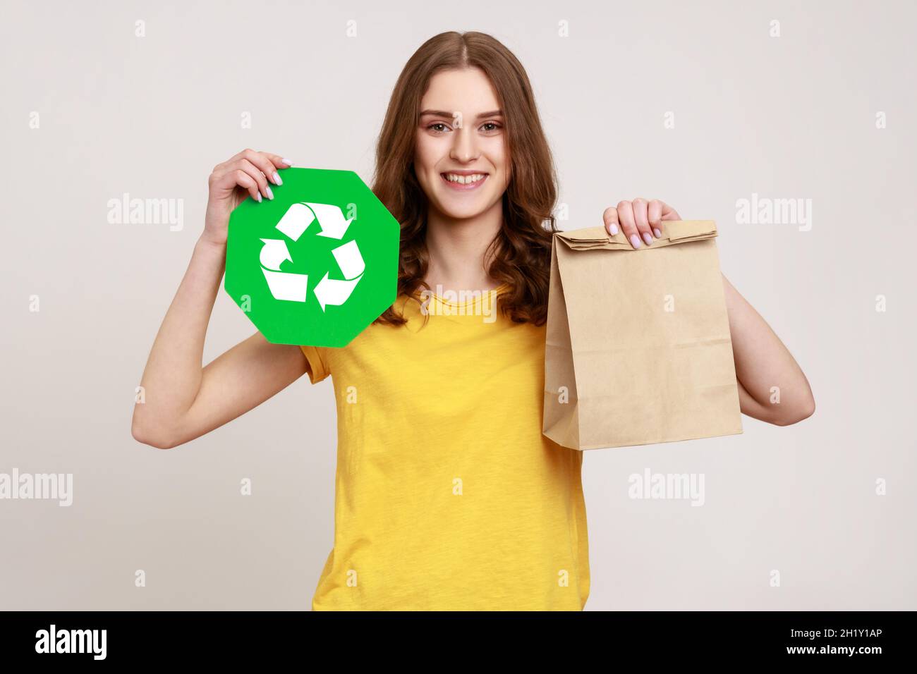 Lächelndes Teenager-Mädchen in gelbem legerem T-Shirt mit Papierpaket und grünem Recycling-Schild, mit Blick auf die Kamera, mit dem Ausdruck von Glück. Innenaufnahme des Studios isoliert auf grauem Hintergrund. Stockfoto