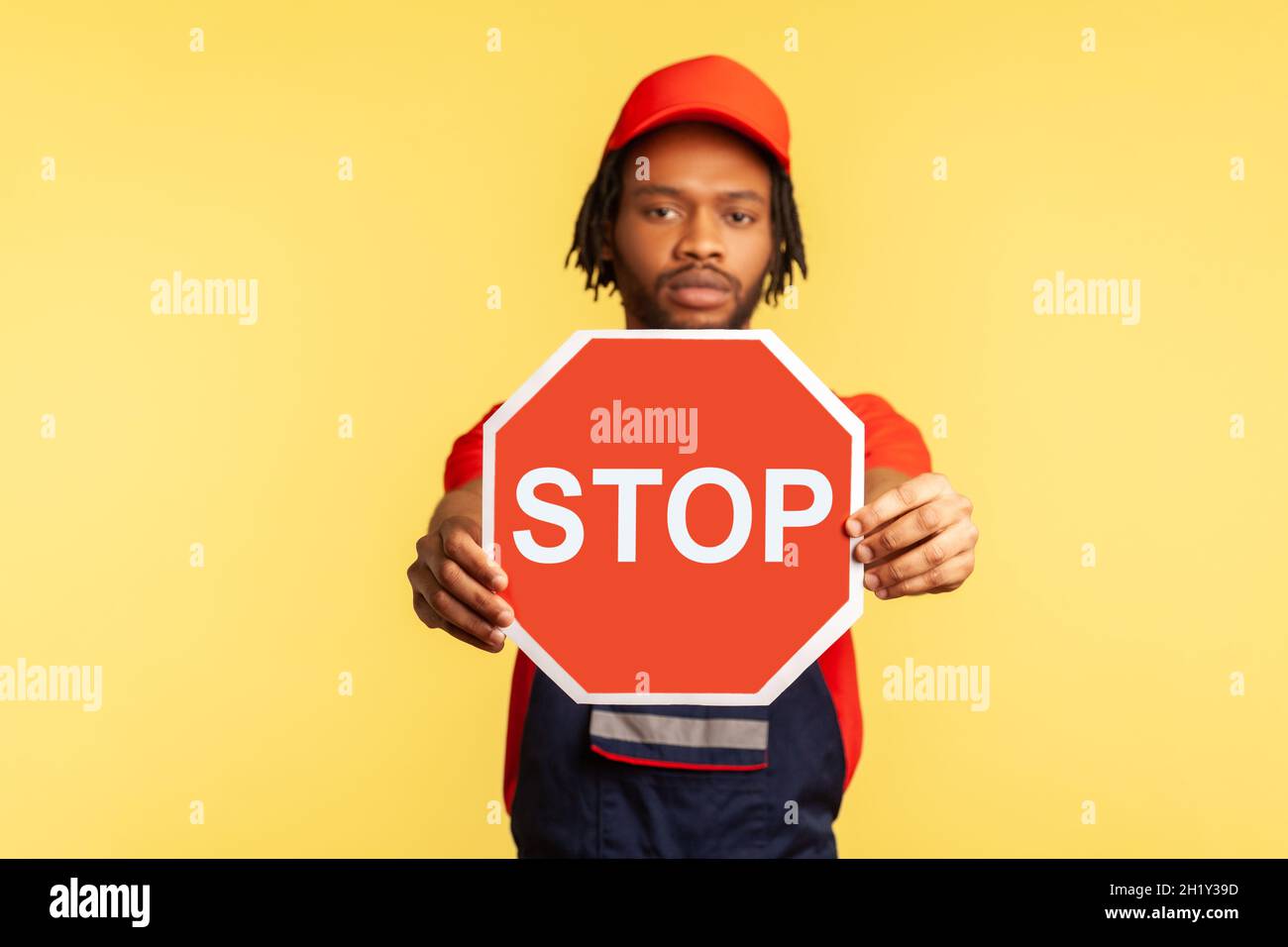 Porträt eines ernsthaften Handwerkers, der Uniform und rote Visierkappe trägt und die Kamera anschaut, mit einem roten Stoppschild, warnt vor Gefahren. Innenaufnahme des Studios isoliert auf gelbem Hintergrund. Stockfoto
