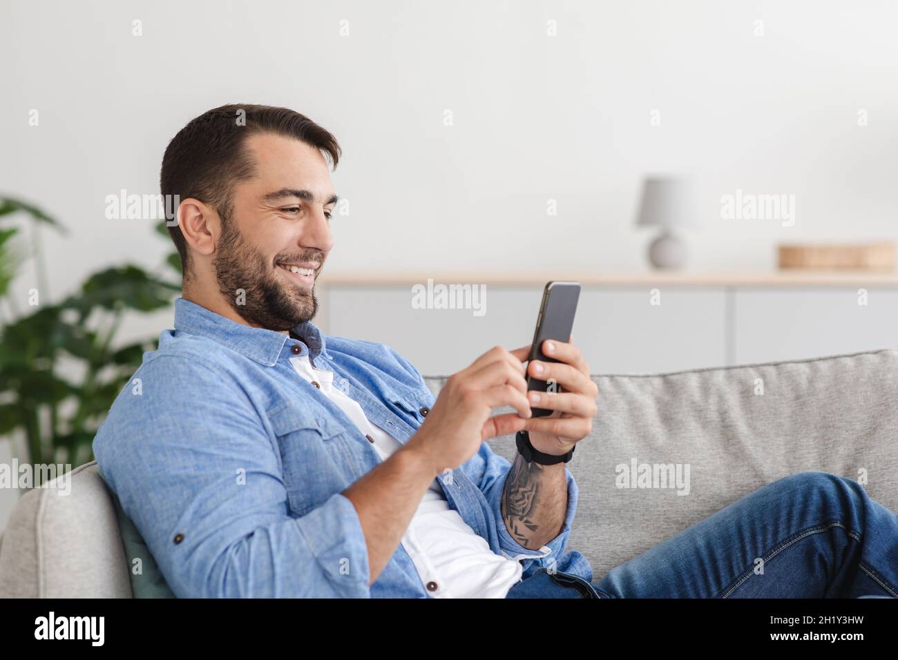 Froher, erwachsener Kaukasusmann mit Bart auf dem Sofa sitzend und auf dem Smartphone chattet Stockfoto