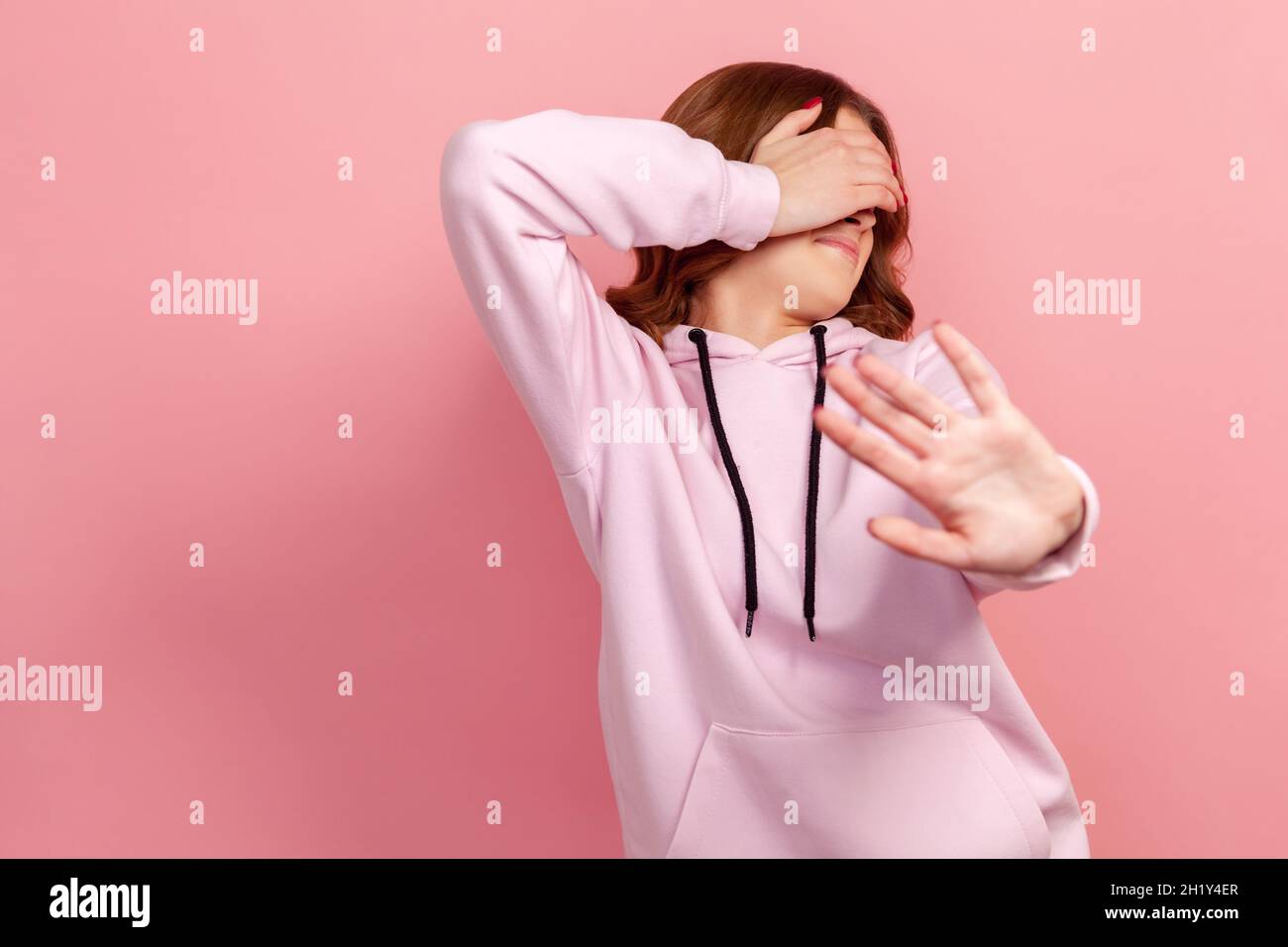 Porträt einer ängstlichen oder schüchternen jungen Frau mit lockigem Haar im Hoodie, die die Augen mit der Handfläche schließt und eine Stop-Hand-Geste zeigt. Innenaufnahme des Studios isoliert auf rosa Hintergrund Stockfoto