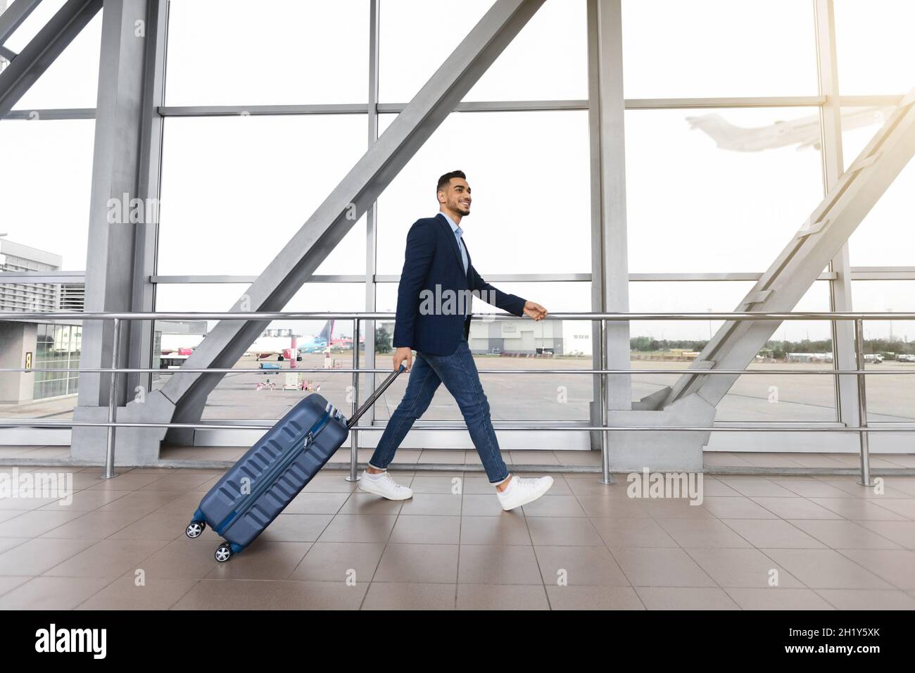 Porträt Eines Lächelnden Arabischen Geschäftsmannes Mit Koffer, Der In Der Nähe Von Window Am Flughafen Herumläuft Stockfoto