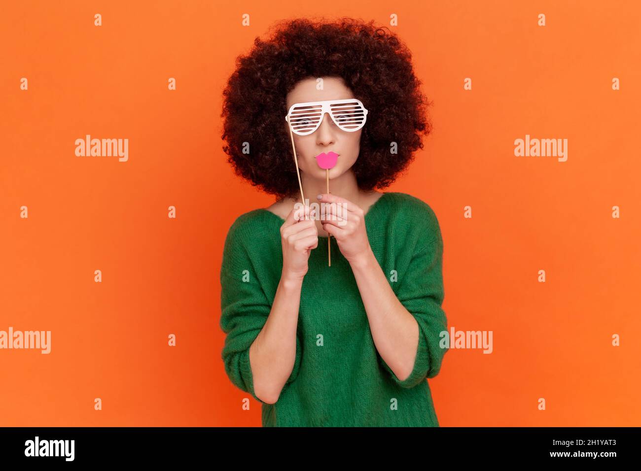Lustige Frau mit Afro-Frisur trägt einen grünen Pullover im Casual-Stil, der die Augen mit einer Papierbrille bedeckt, rote Lippen auf dem Stock hält und sich maskiert. Innenaufnahme des Studios isoliert auf orangefarbenem Hintergrund. Stockfoto