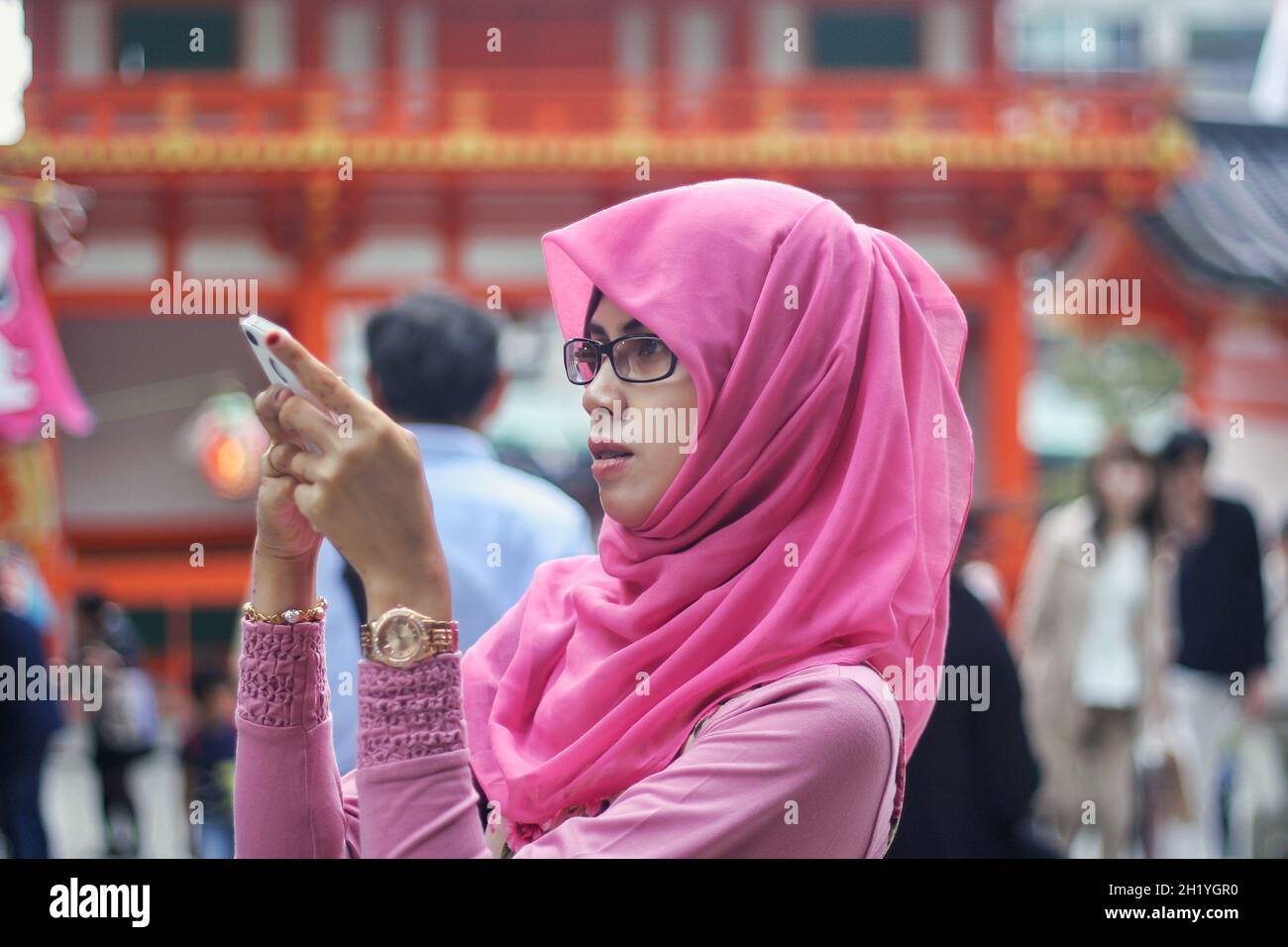 Nahaufnahme eines Portraits einer jungen asiatischen muslimischen Frau mit pinkfarbenem Hijab und einer Brille, die ein Smartphone mit Bokeh-Hintergrund auf der Straße hält und anschaut. Stockfoto