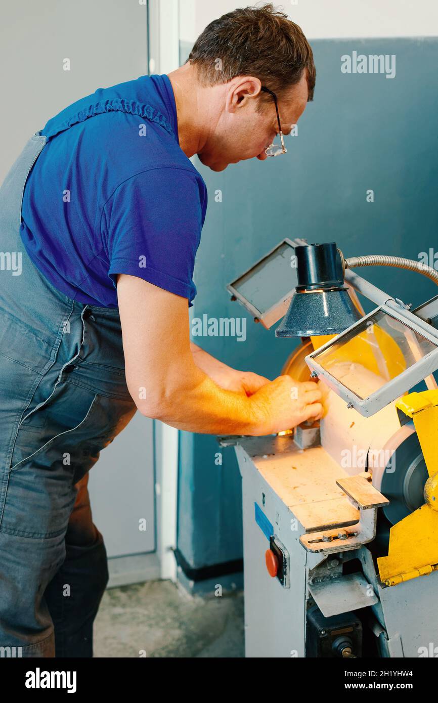 Ein kaukasischer Arbeiter in Uniform schärft in einer Werkstatt ein Teil an einer Maschine. Manuelle Verarbeitung von Metall. Ein wahres Porträt eines Arbeiters. Stockfoto