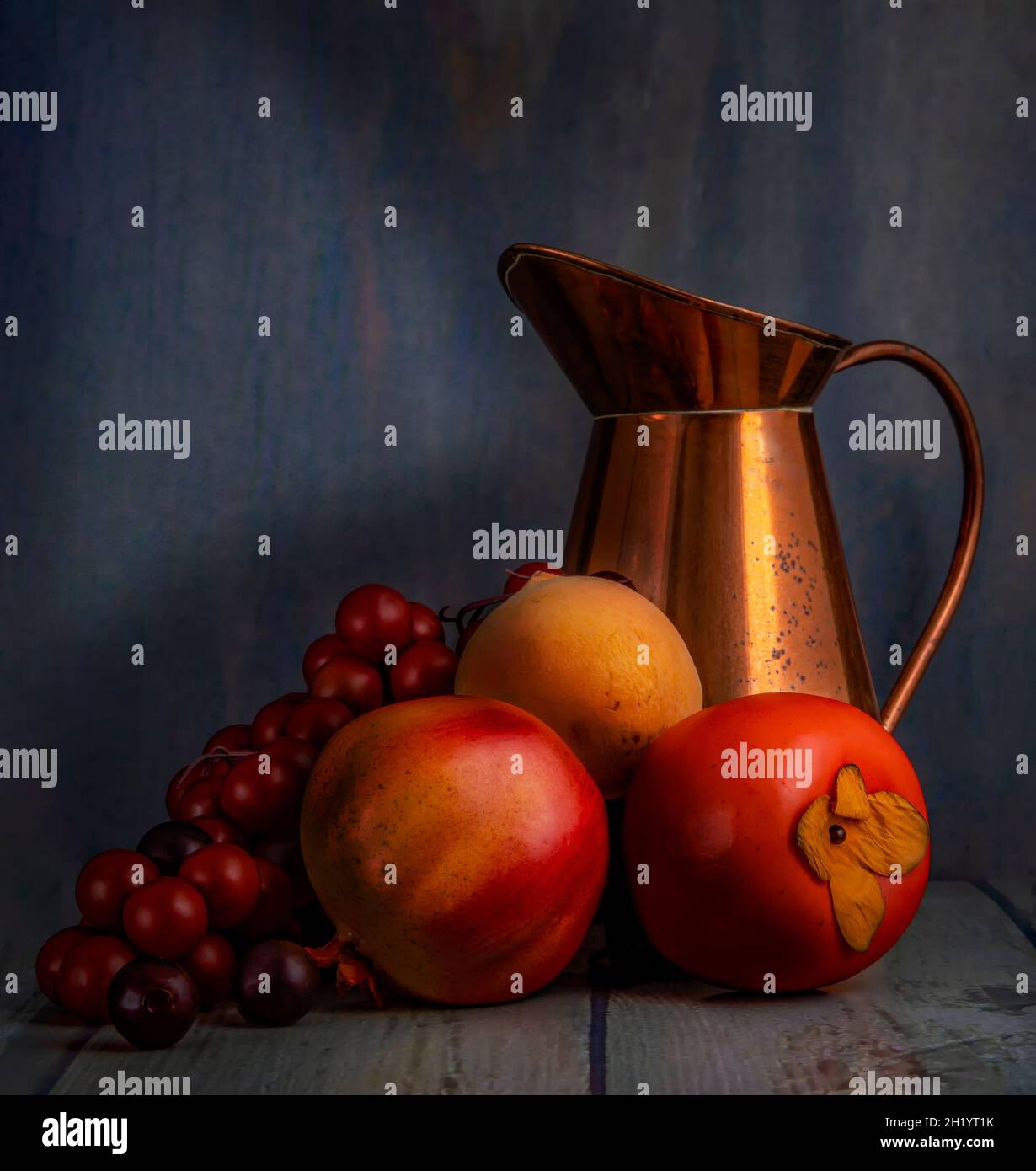 Künstlerische Stillleben fotografieren rote Trauben. Zitrone, Kaki und Messingkrug auf einem Holztisch, mit dunklem Hintergrund. Stockfoto