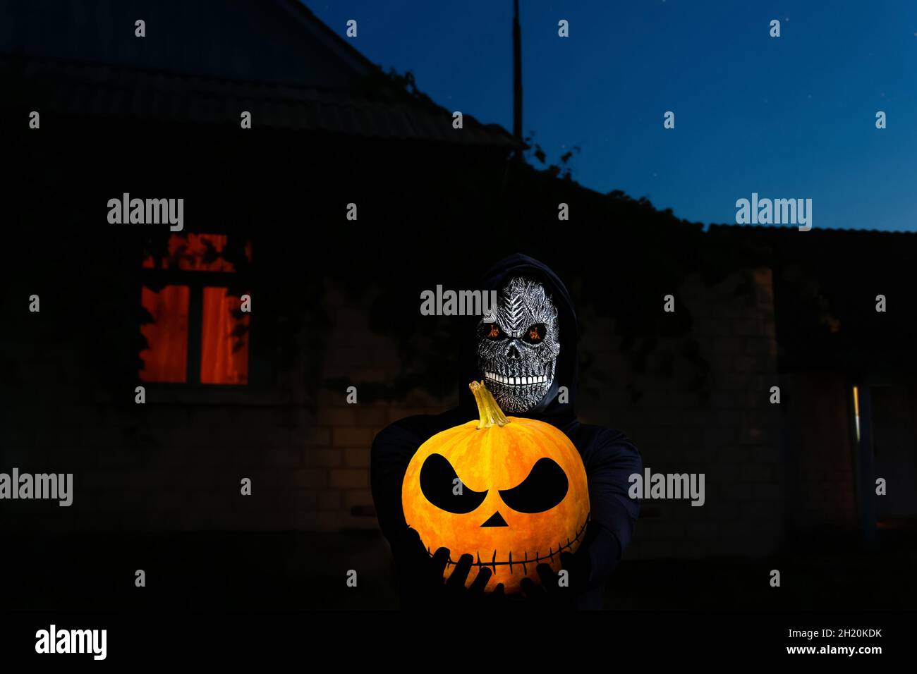 Grimmiger Schnitter hält halloween Kürbis Kopf. Mann in Totenmaske mit Feuerflamme in den Augen am Nachthimmel, heller Fensterhintergrund. Halloween Feiertagskonzept Stockfoto